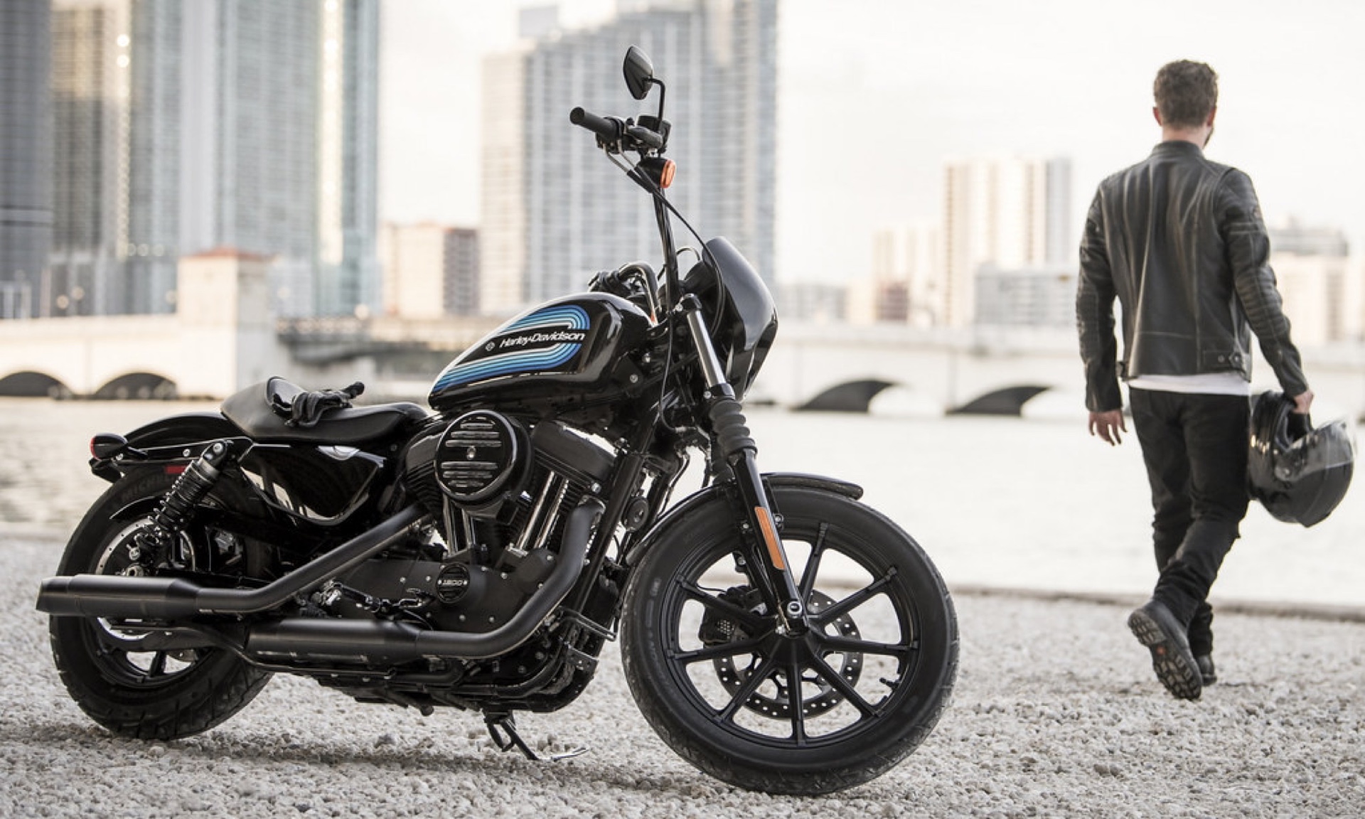 Harley-Davidson motorcycle with a rider carrying a helmet beside a city skyline.